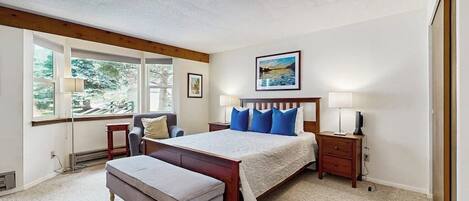 A bedroom with a large bed, a bench, a nightstand, a lamp, and a window with a view of greenery.