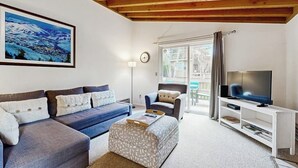 Un salon comprenant un canapé, un pouf, un meuble pour la télévision et un tableau représentant un paysage enneigé.