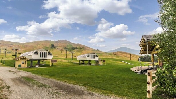 Un cadre rural avec deux cabanes blanches à un seul étage, dotées de v érandas attenantes, et entourées de champs verdoyants et de collines.