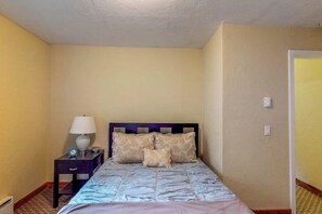 Een slaapkamer met een bed, een nachtkastje met een lamp en een aan de muur bevestigde thermostaat.