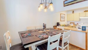 A dining area with a wooden table, white chairs, a chandelier, and a kitchen in the background. - Newly updated condo - close to downtown Ketchum & the ski lifts (Ketchum)