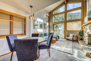 A spacious living area with a round table, multiple seating options, large windows, and a stone fireplace.