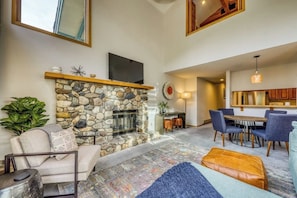 A living room with a stone fireplace, a television, a dining area with blue chairs, and a kitchen in the background.
