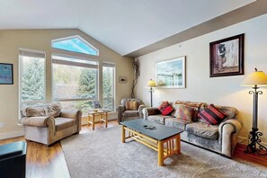 A living room with two sofas, a coffee table, and a floor lamp. There are large windows and a framed picture on the wall.