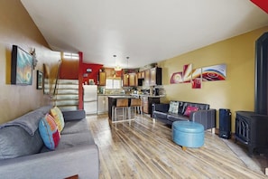 Ein Wohnzimmer mit grauem Sofa, blauem Hocker, einer Küchenzeile und einem schwarzen Kaminofen.