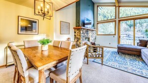 A dining area with a wooden table and chairs, a stone fireplace, and large windows with a view of trees. - Newly-updated Warm Springs condo w/ski & mountain views - Dogs ok (Ketchum)