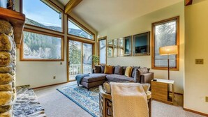 A cozy living room with a sectional sofa, a small table, and a lamp. Large windows offer a view of the outdoors. - Newly-updated Warm Springs condo w/ski & mountain views - Dogs ok (Ketchum)