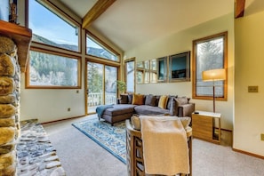 A cozy living room with a sectional sofa, a small table, and a lamp. Large windows offer a view of the outdoors.