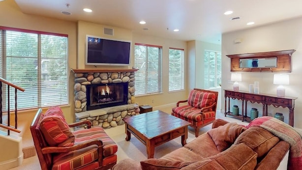 A living room with a stone fireplace, a television mounted above it, and a wooden coffee table. - Comfortable, centrally-located condo close to skiing, hiking, & downtown (Ketchum)