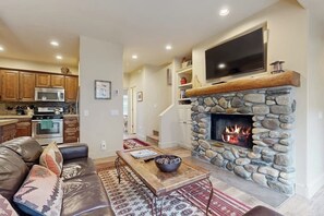 Ein Wohnzimmer mit Kamin aus Stein, einem Flachbildfernseher, einem Ledersofa und einer Küche im Hintergrund.