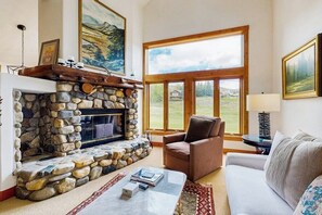 Ein Kamin aus Stein, ein brauner Ledersessel, ein weißes Sofa und ein großes Fenster mit Blick auf eine grüne Landschaft.