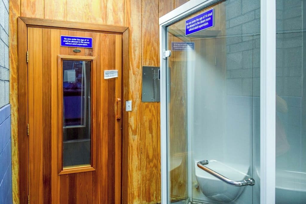 A wooden door with a sign indicating no one under 14 without adult supervision, and a glass shower stall with a handrail.