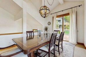Ein Esszimmer mit einem großen Holztisch, Stühlen, einer Chandelier und Blick auf ein Haus durch ein Fenster.