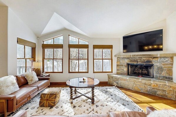 A cozy living room with a stone fireplace, a large TV mounted above it, and a high ceiling with skylights. - Spacious mountain getaway near the slopes with riverfront view & garage (Ketchum Idaho)