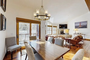 A spacious dining area with a large wooden table, several chairs, a chandelier, and a television on the wall. - Spacious mountain getaway near the slopes with riverfront view & garage (Ketchum Idaho)