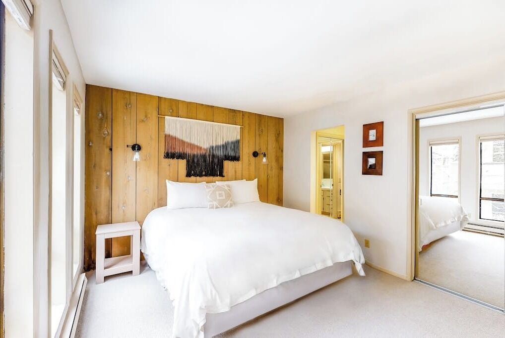 A bedroom with a wooden headboard, a bed with white bedding, a small bedside table, and a mirror on the wall.