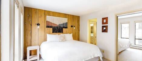 A bedroom with a wooden headboard, a bed with white bedding, a small bedside table, and a mirror on the wall.
