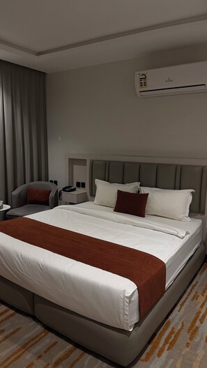 A hotel room with a large bed, a chair, a bedside table, and an air conditioning unit.