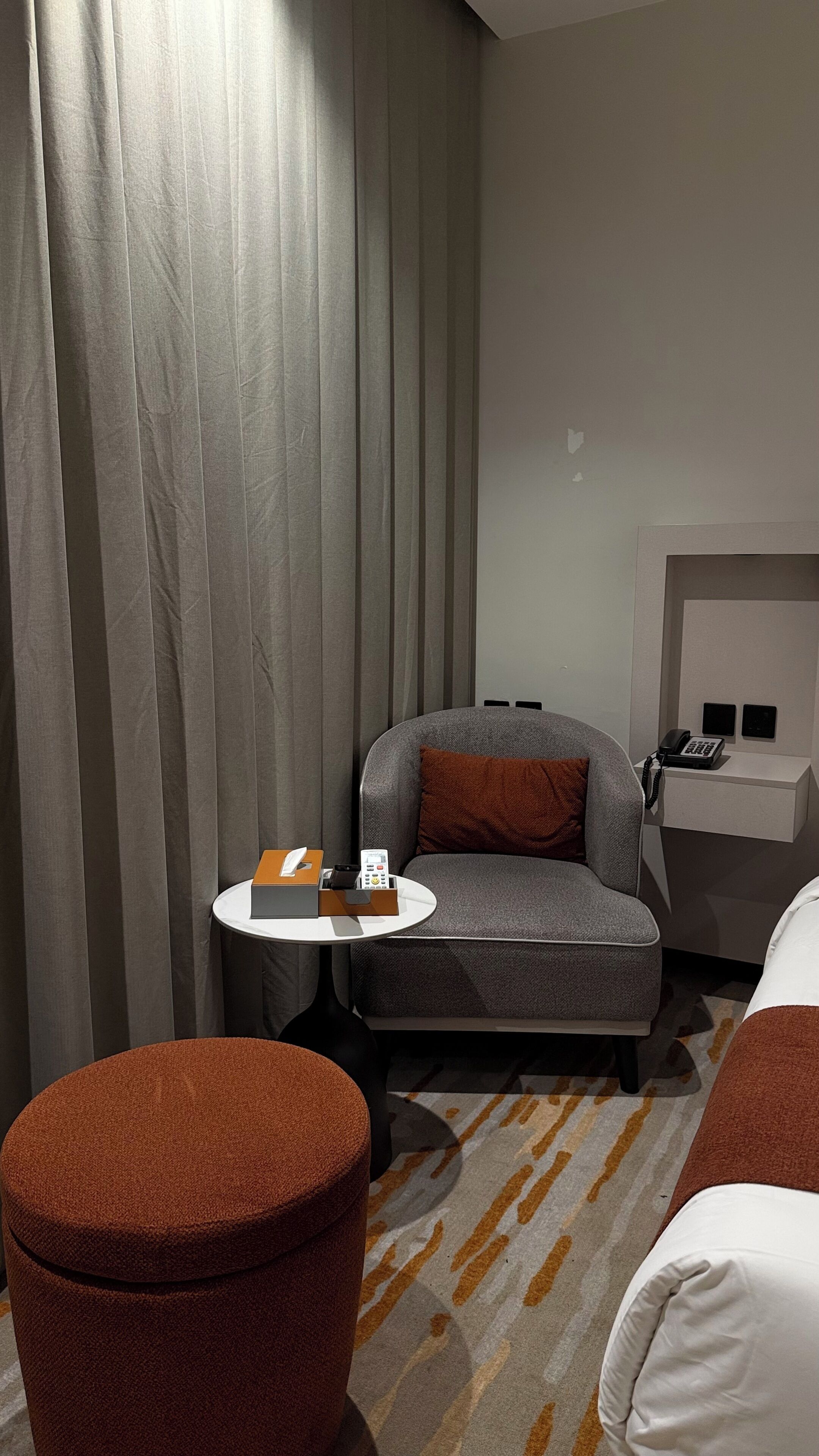 Moderni hotellihuone, jossa on harmaa sohva, pieni pyöreä pöytä, jossa on puhelin ja rasia, ruskea jalkarahi ja valkoinen sänky.