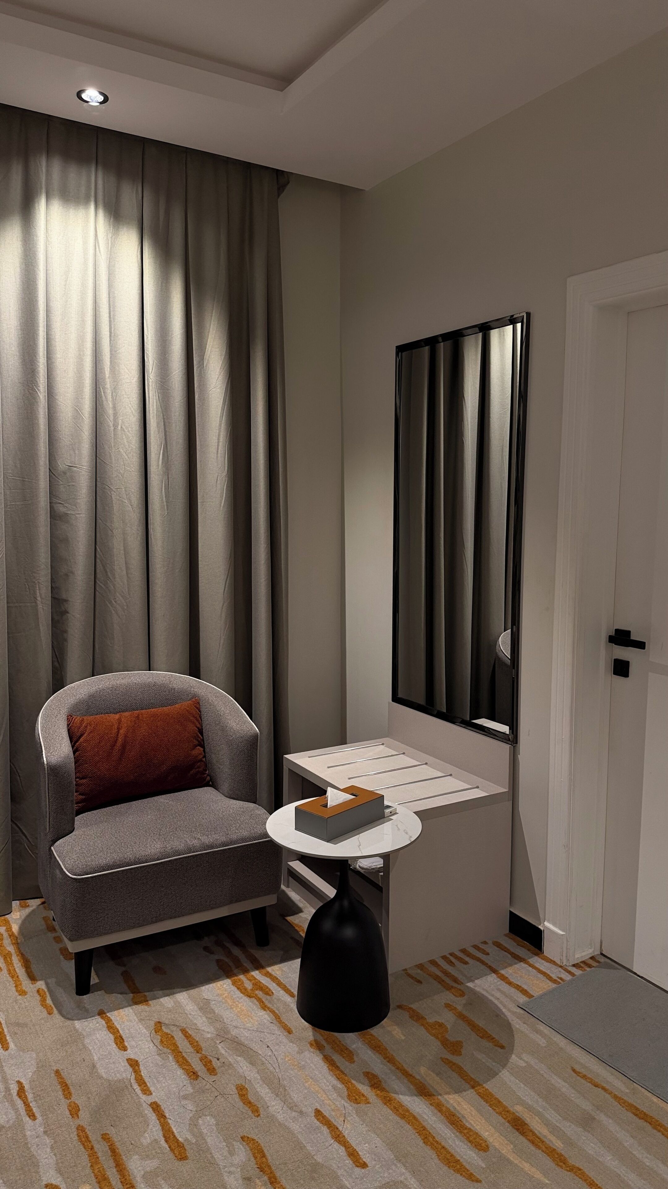 Une chambre moderne avec un fauteuil gris, une petite table ronde sur laquelle se trouve une boîte de mouchoirs, un grand miroir et une porte.