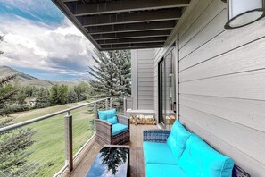 Ein Balkon mit einer Couch, Korbstühlen, einer Glastür und Blick auf eine gebirgige Landschaft.