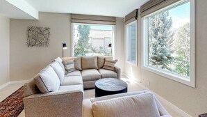 Ein modernes Wohnzimmer mit einer Eckcouch, einem runden Couchtisch und großen Fenstern mit Blick auf Bäume.