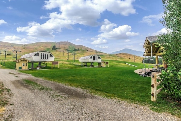 A rural setting with two white, semi-circular buildings with attached garages, surrounded by green fields and hills. - Golf course condo with ski shuttle, shared pools, & hot tub (Sun Valley)