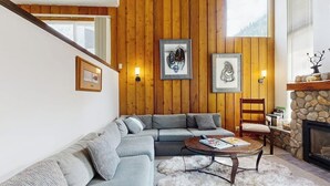 A living room with a sectional sofa, a round coffee table, a stone fireplace, and wooden panel walls. - Wonderful Mountain Getaway W/ Shared Pool, Multiple Balconies, Gas Grill & WiFi (Ketchum)