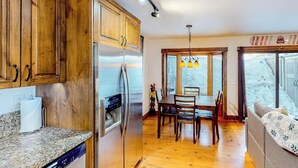 Una cocina con armarios de madera, un refrigerador de acero inoxidable, una mesa de comedor con sillas y un ventanal con vistas a un paisaje nevado.