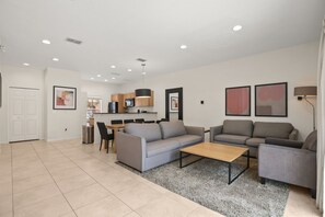 Ein modernes Wohnzimmer mit grauem Sofa, einem Couchtisch und einem grauen Sessel. Es gibt eine Küchenzeile mit Holzschränken und einem Esstisch mit Stühlen.