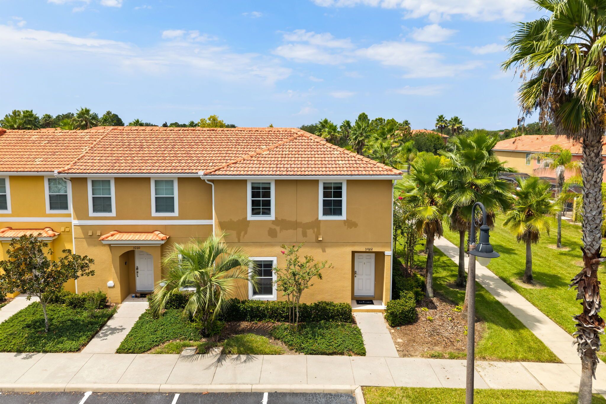 A two-story residential building with a tiled roof, a front entrance, and a small garden area with palm trees and shrubs.