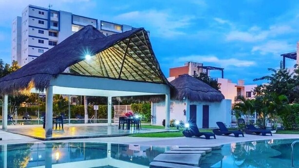 A swimming pool with lounge chairs, a thatched-roof structure, and modern buildings in the background. - Modern 2BR 2BA | Workspace Wifi | Pools | 24/7 Sec (Cancún)