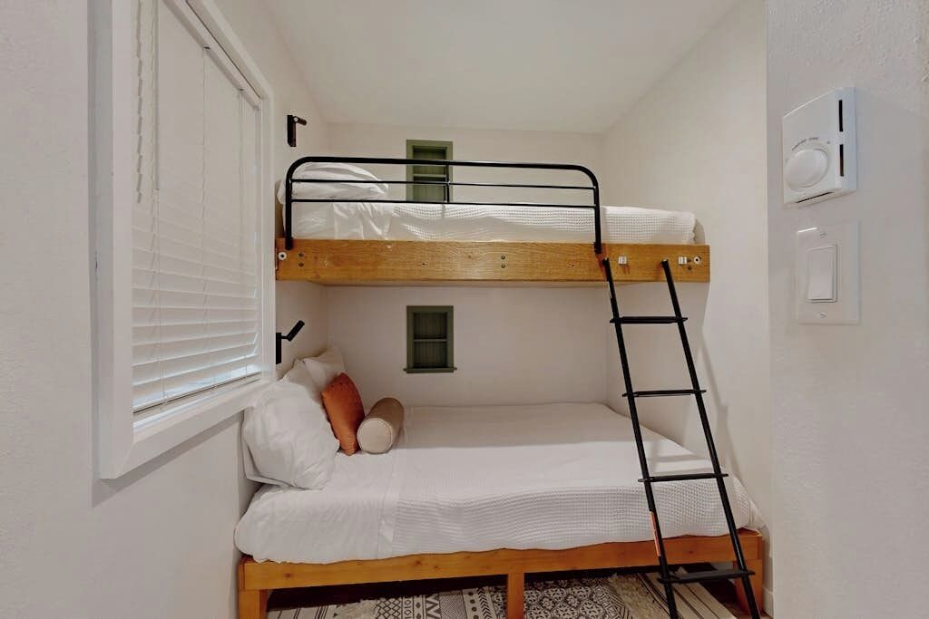 Een stapelbed met een ladder, een raam met zonwering en een aan de muur bevestigde thermostaat.
