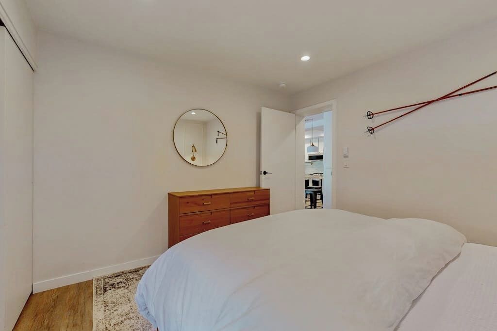 Un dormitorio con cama, cómoda de madera, espejo redondo y una puerta que da a otra habitación.