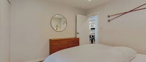 Un dormitorio con cama, cómoda de madera, espejo redondo y una puerta que da a otra habitación.