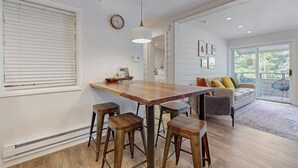 Une cuisine moderne avec une table en bois et des tabourets de bar, un luminaire suspendu et une vue sur un espace de vie comprenant un canapé et des fauteuils.