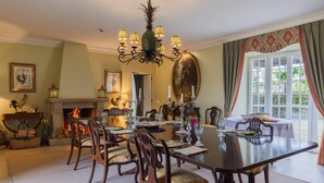 Une salle à manger avec une grande table, des chaises, un lustre, une cheminée et une grande fenêtre avec des rideaux.
