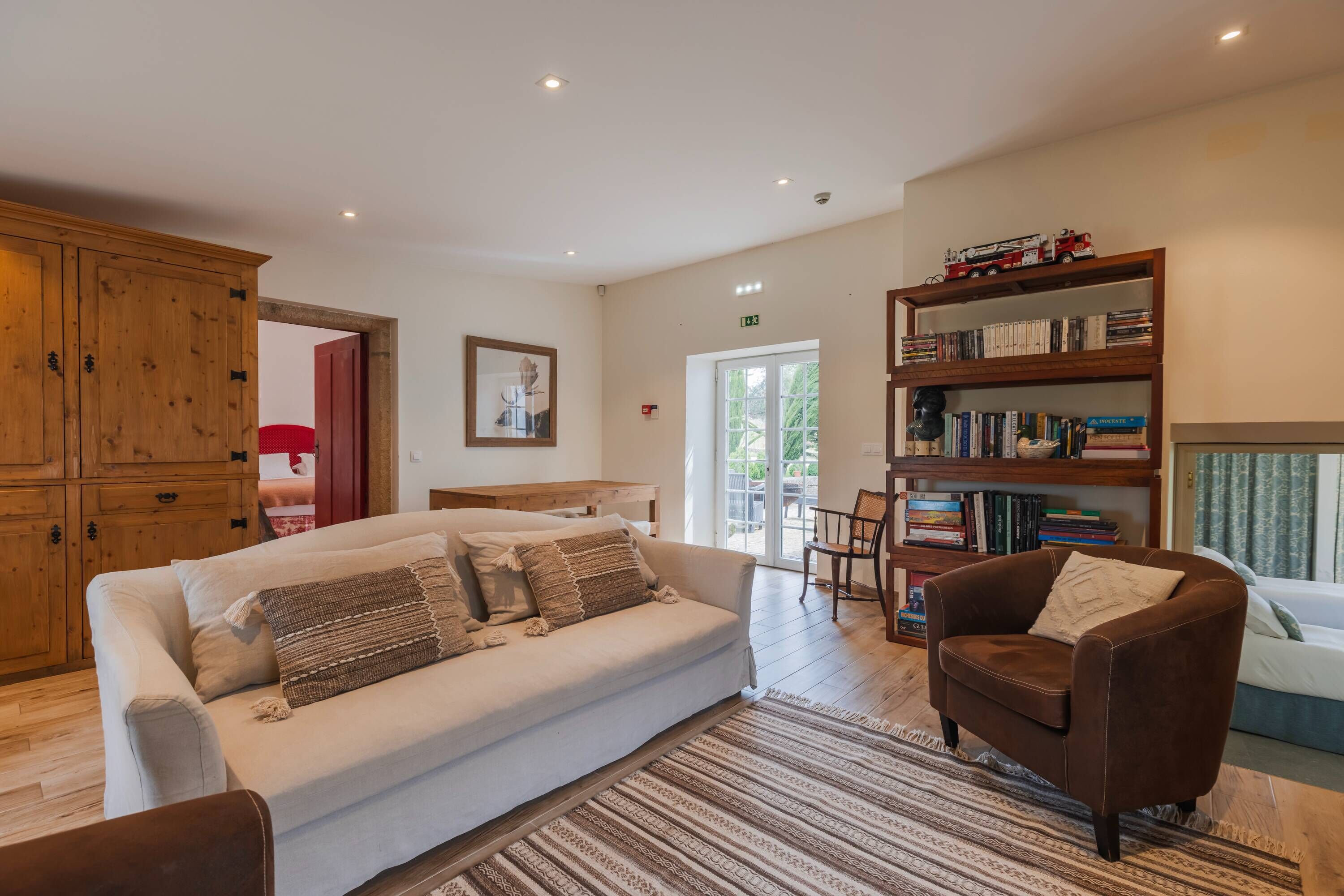 A spacious living area with a sofa, armchair, wooden cabinet, bookshelf, and a door leading to a balcony.