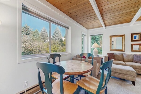 A room with a round table, chairs, a sofa, and a large window with a view of trees.