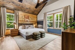 Ein Schlafzimmer mit Holzboden, einem Bett mit Kissen, einem Nachttisch, einem Fenster mit Vorhängen und einer Pflanze.