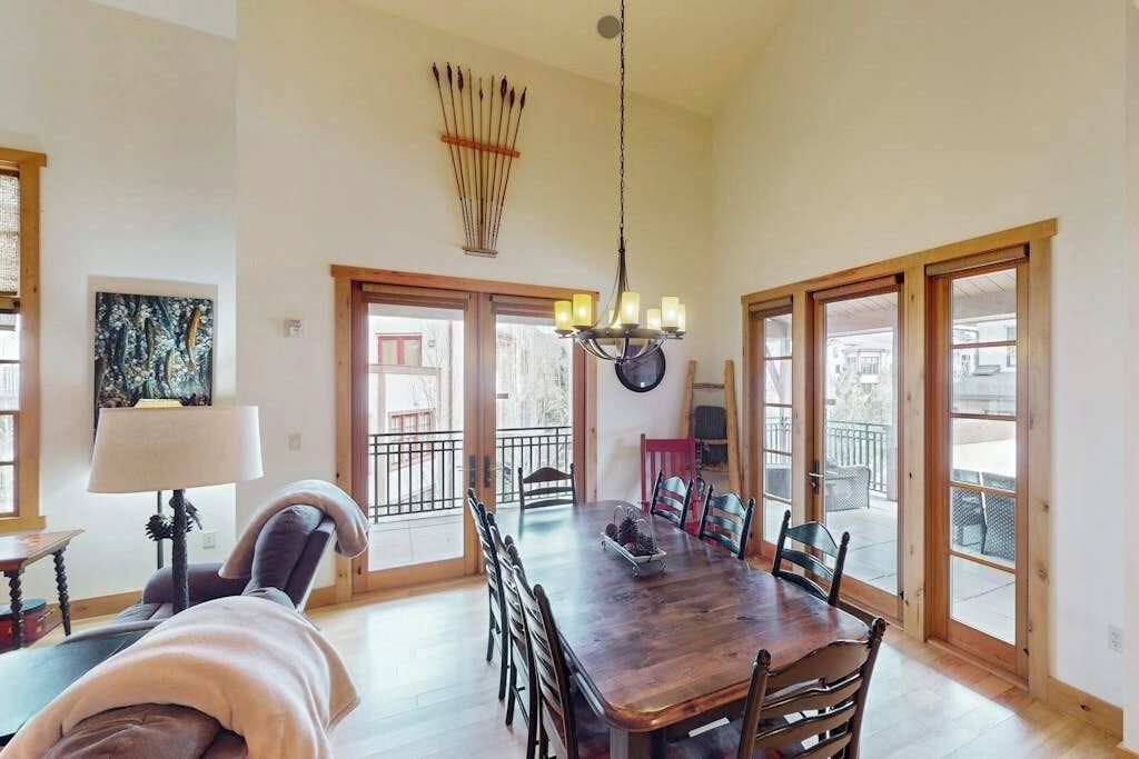 A dining area with a large wooden table, chairs, a chandelier, and large windows with a view.