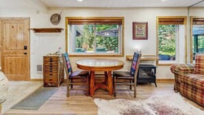 Ein Holztisch mit Stühlen, eine Couch, eine Kommode und ein Kuhfellteppich in einem Raum mit Holzbalken und großen Fenstern.