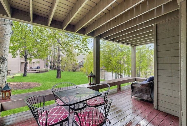 A covered outdoor patio with a table and chairs, a wooden deck, and a view of a green lawn and trees.