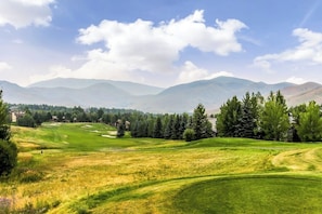Ein Golfplatz mit sanften Hügeln, Bäumen und fernen Bergen unter blauem Himmel.
