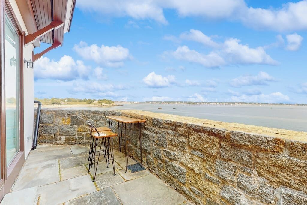 En stenvæg med udsigt til havet, et træbord og stole på en terrasse.