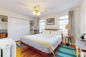 Ein Schlafzimmer mit Bett, Deckenventilator, Fenster mit Vorhängen, Nachttisch mit Lampe und Heizkörper.