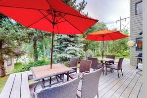 Une terrasse extérieure avec une table en bois et des chaises en osier, deux parasols rouges et une vue sur une zone boisée.