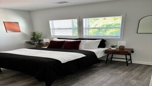 Un dormitorio con una cama, dos ventanas, una mesita de noche, una planta y un espejo colgado en la pared.