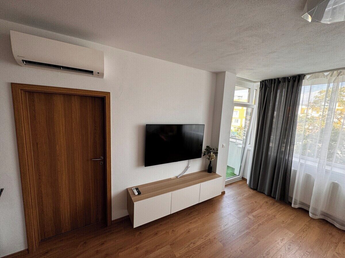 Una habitación con puerta de madera, televisor montado, mesa consola de madera y un amplio ventanal con cortinas.