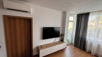 Una habitación con puerta de madera, televisor montado, mesa consola de madera y un amplio ventanal con cortinas.